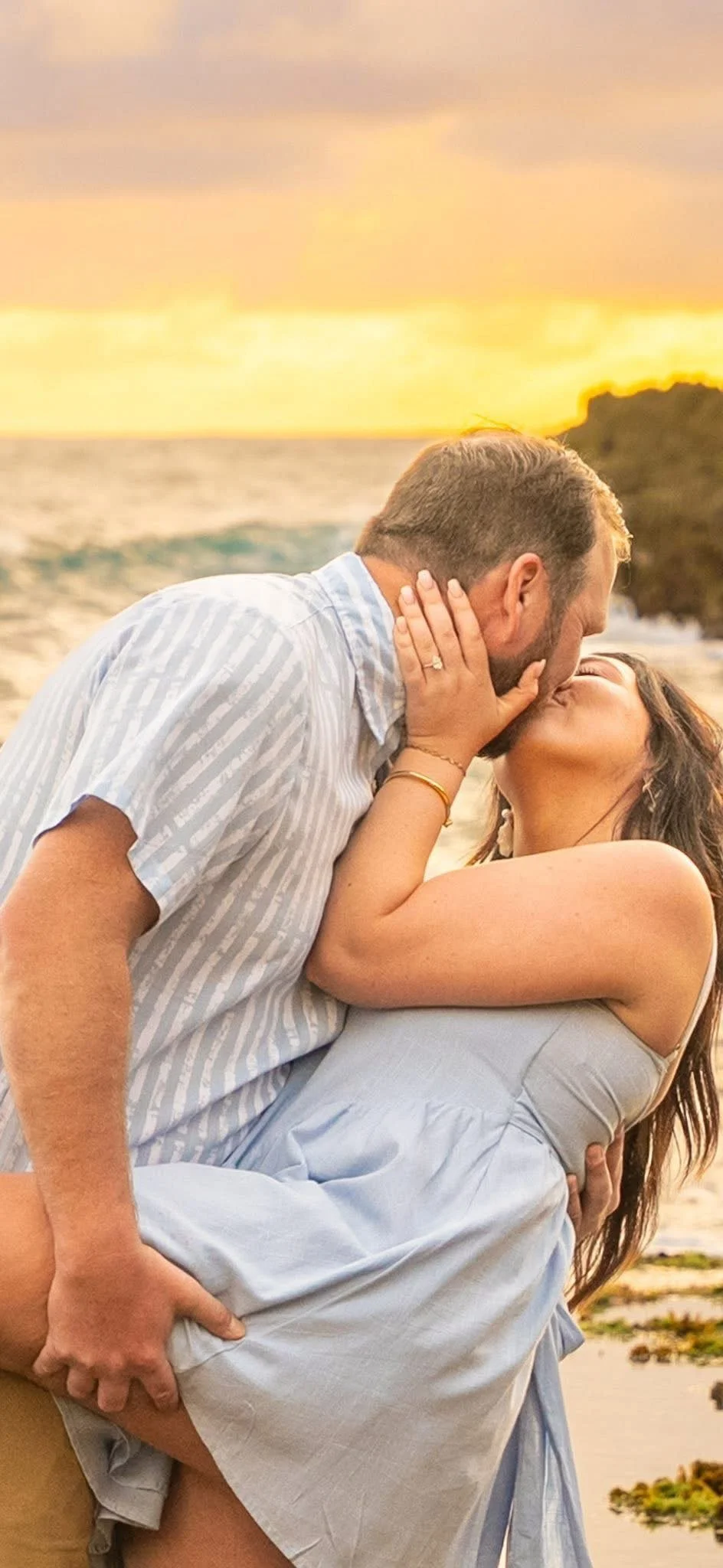 Man lifting his partner for a kiss on the beach at Shipwreck Beach, Kauai, at sunset.