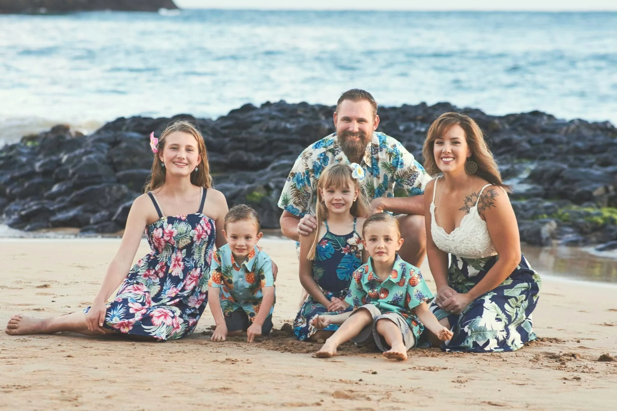 Family of six sits on the sand in front of black lava rocks on a Kauai beach.