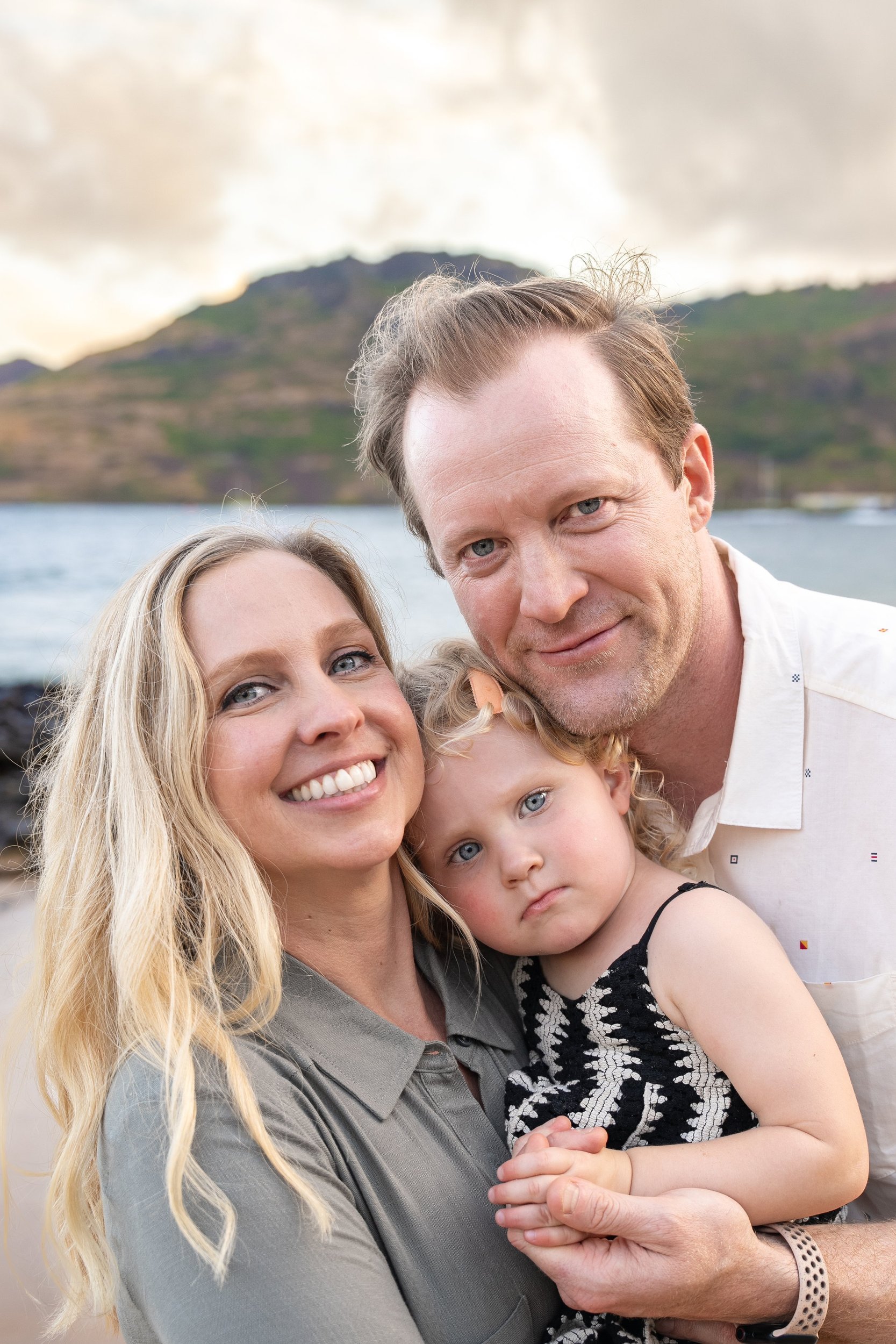 Parents cuddling their toddler daughter close on a Kauai beach with mountains behind them.