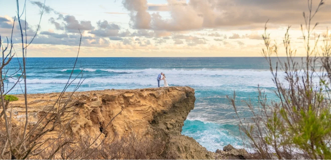 Newlywed couple sharing a kiss on the edge of a dramatic ocean cliff on Kauai, turquoise waves rolling in below a golden sky.