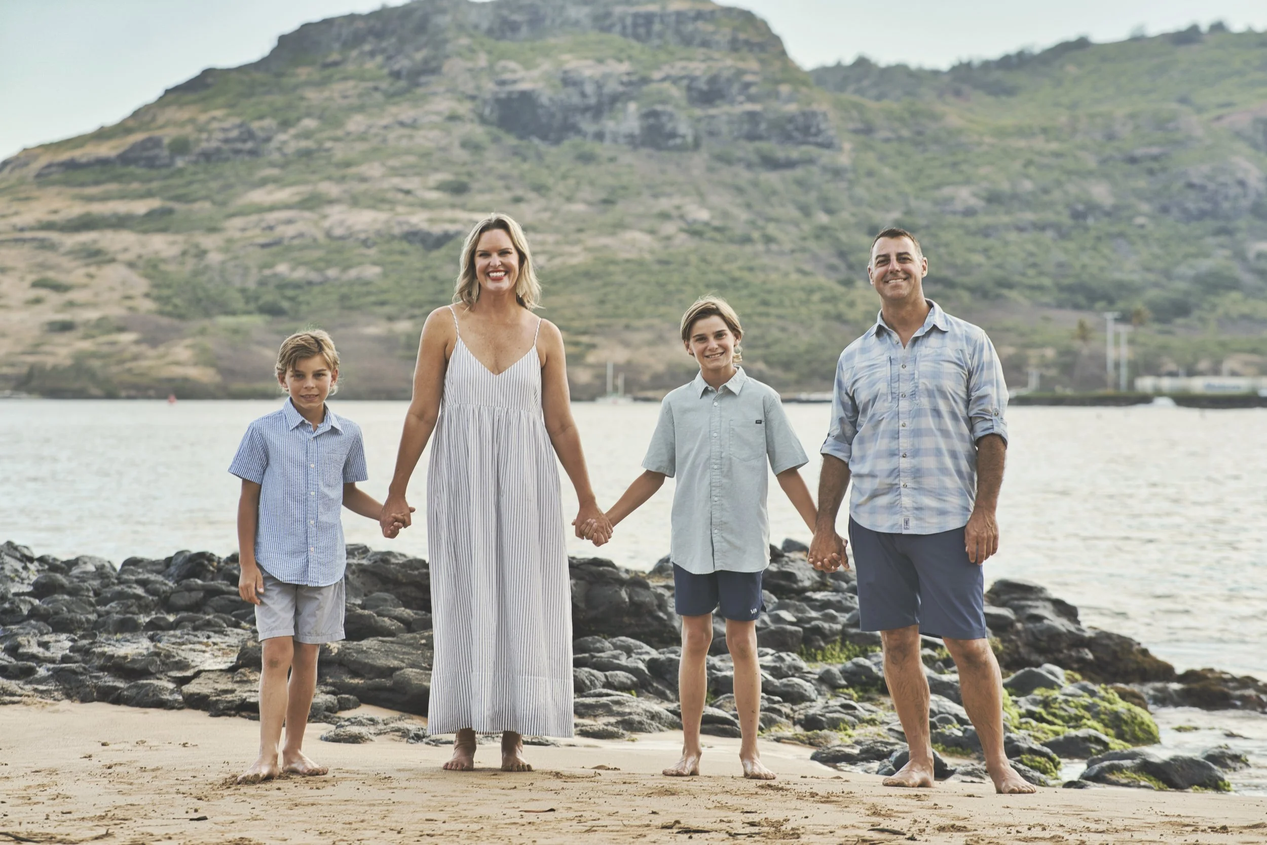 Family of four holding hands on a sandy Kauai beach with green mountains rising behind them.