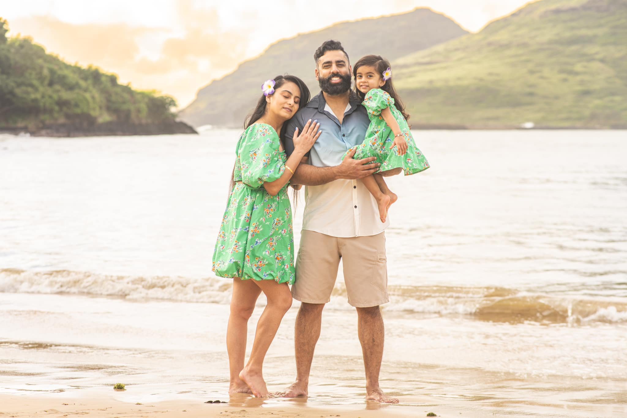 Parents with their toddler daughter on a Kauai beach with green mountains behind them.