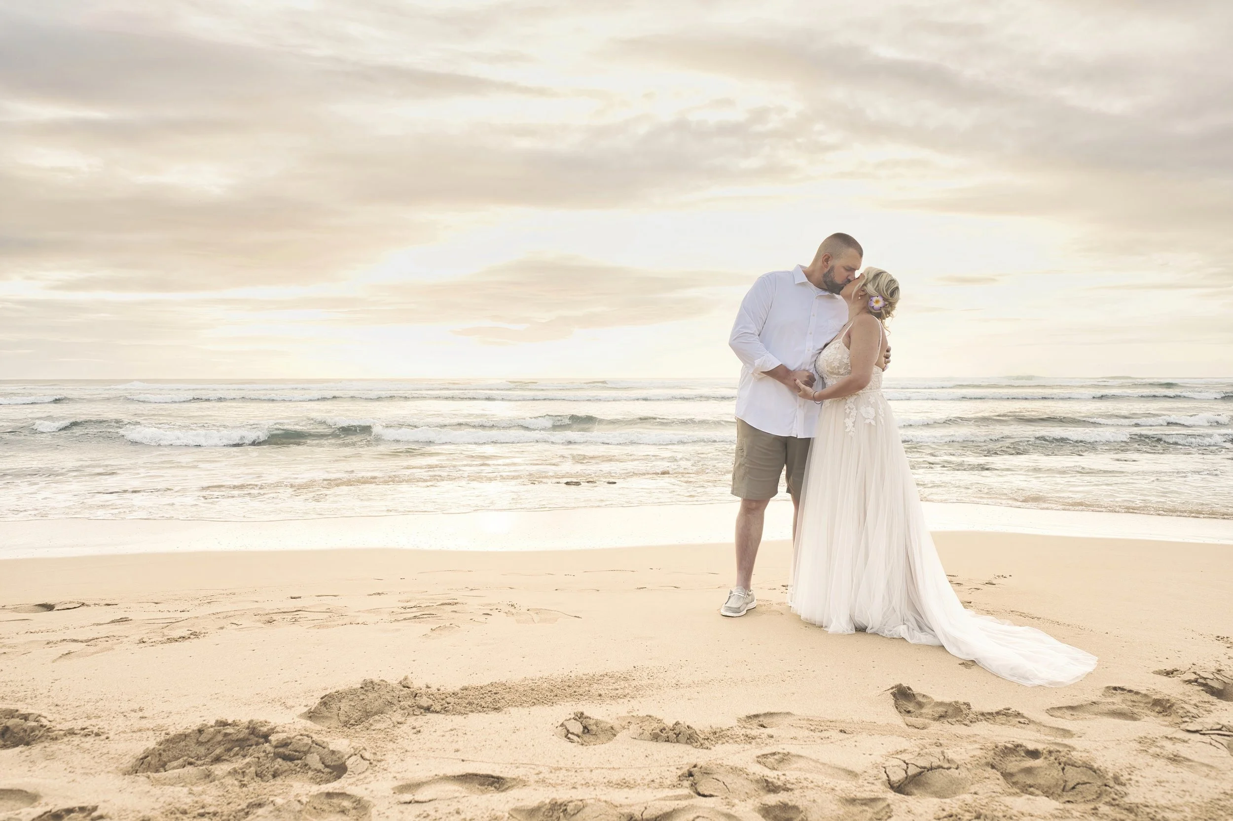 Newlywed couple kissing on a wide sandy Kauai beach at sunset.