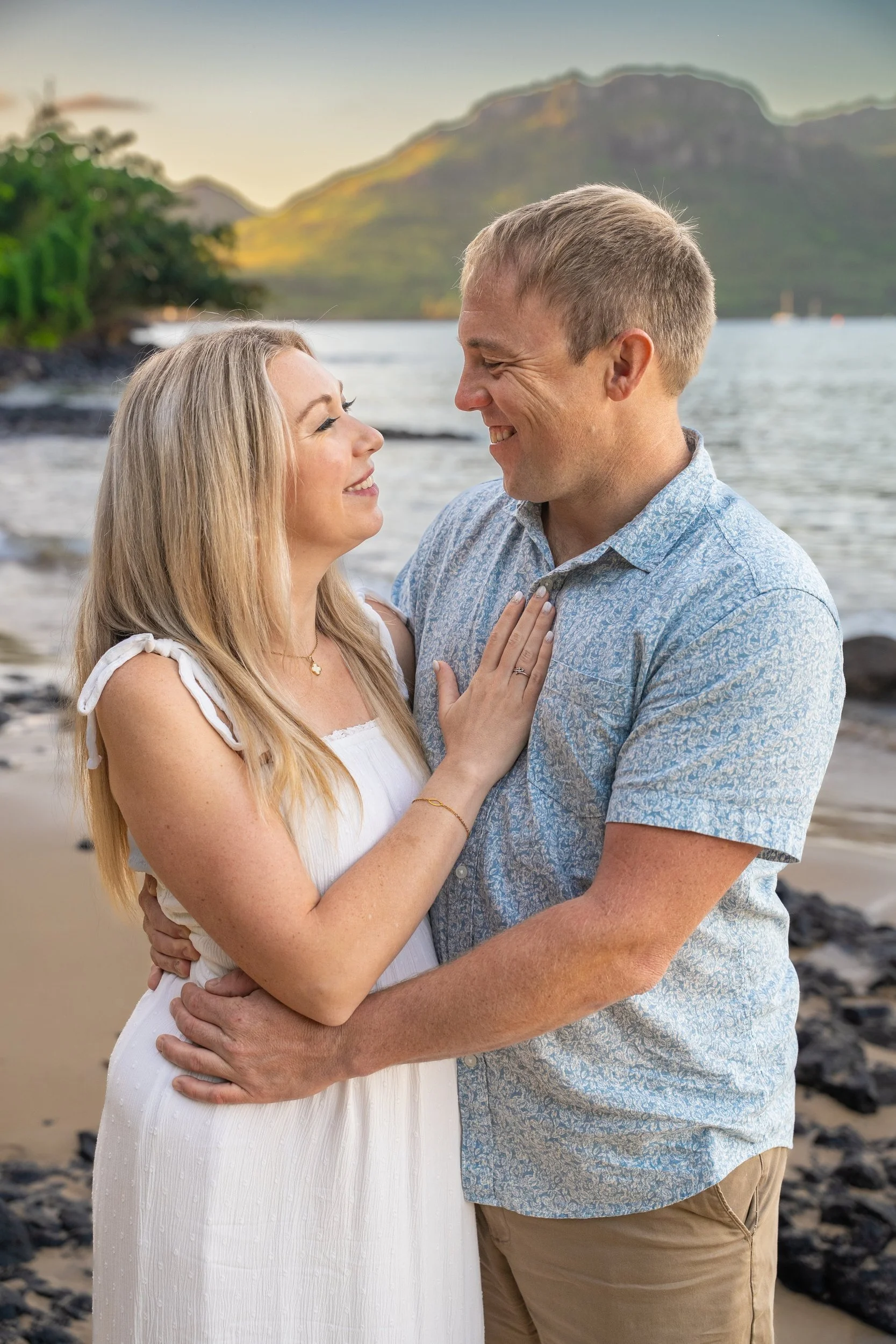 Smiling couple embracing on a Kauai beach with a mountain behind them.