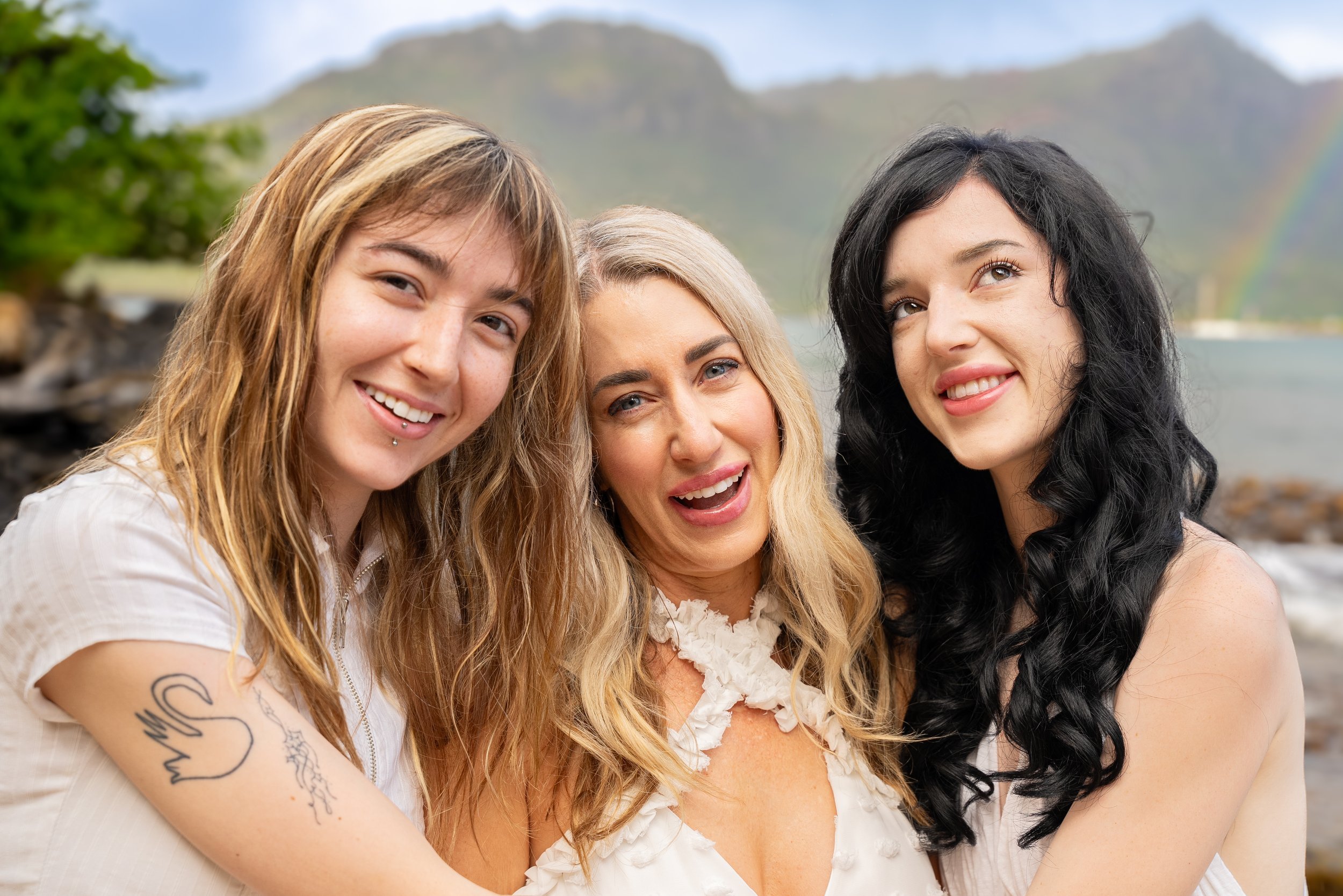 Mom posing with her two adult daughters on a Kauai beach with a rainbow behind them.