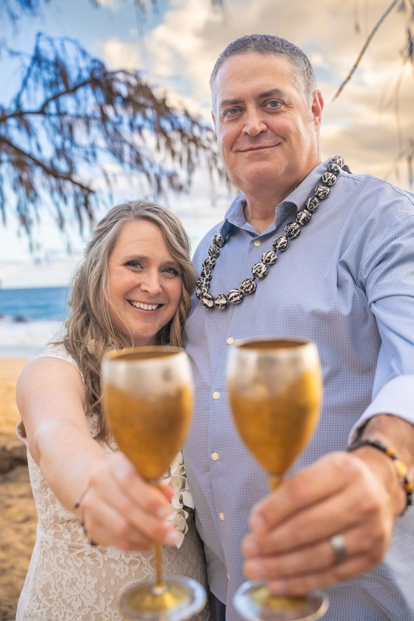 Smiling bride and groom toasting the camera with gold champagne glasses on a Kauai beach, the groom wearing a kukui nut lei.