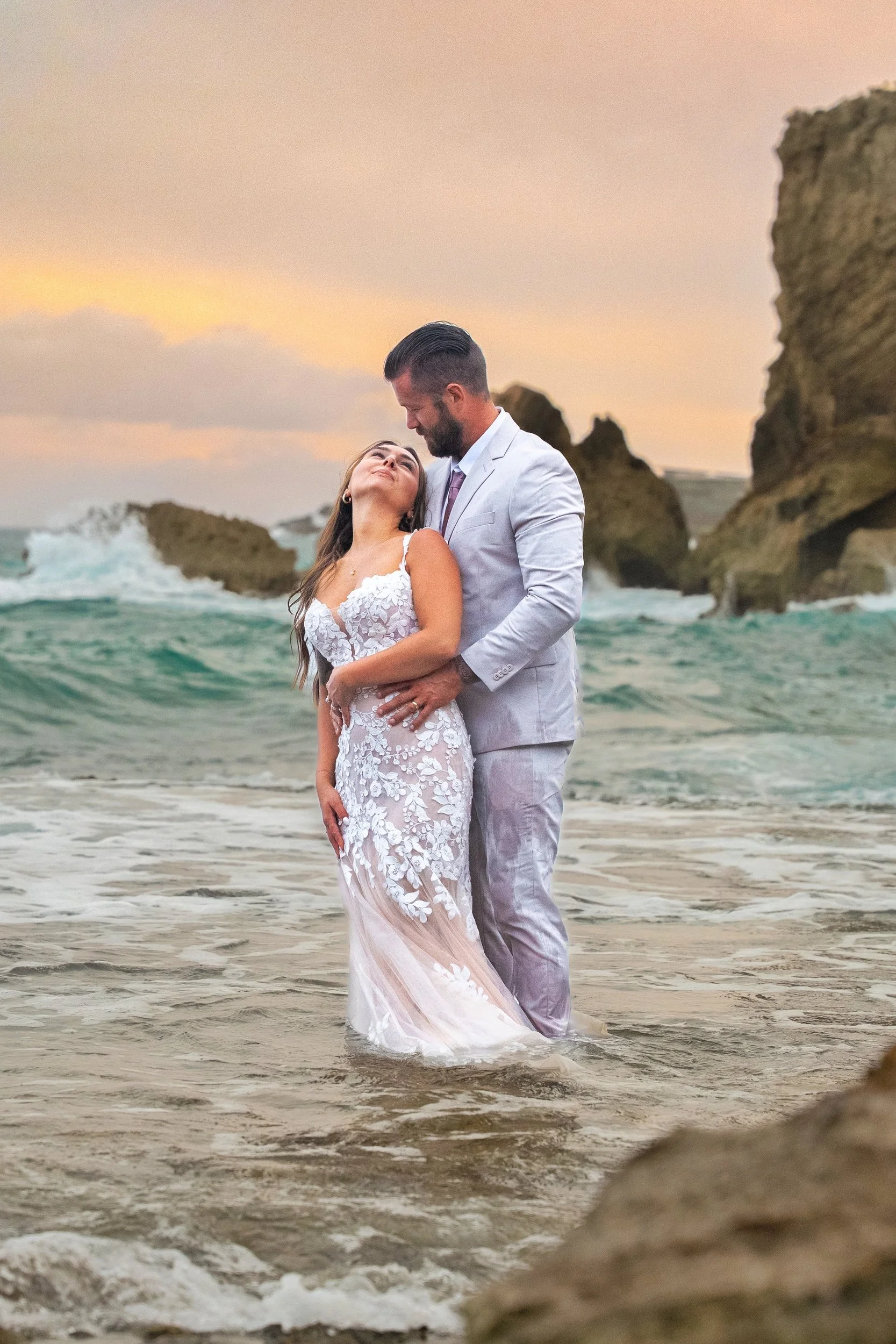 Newlywed couple embracing in the ocean surf beneath sea cliffs on Kauai at sunset.