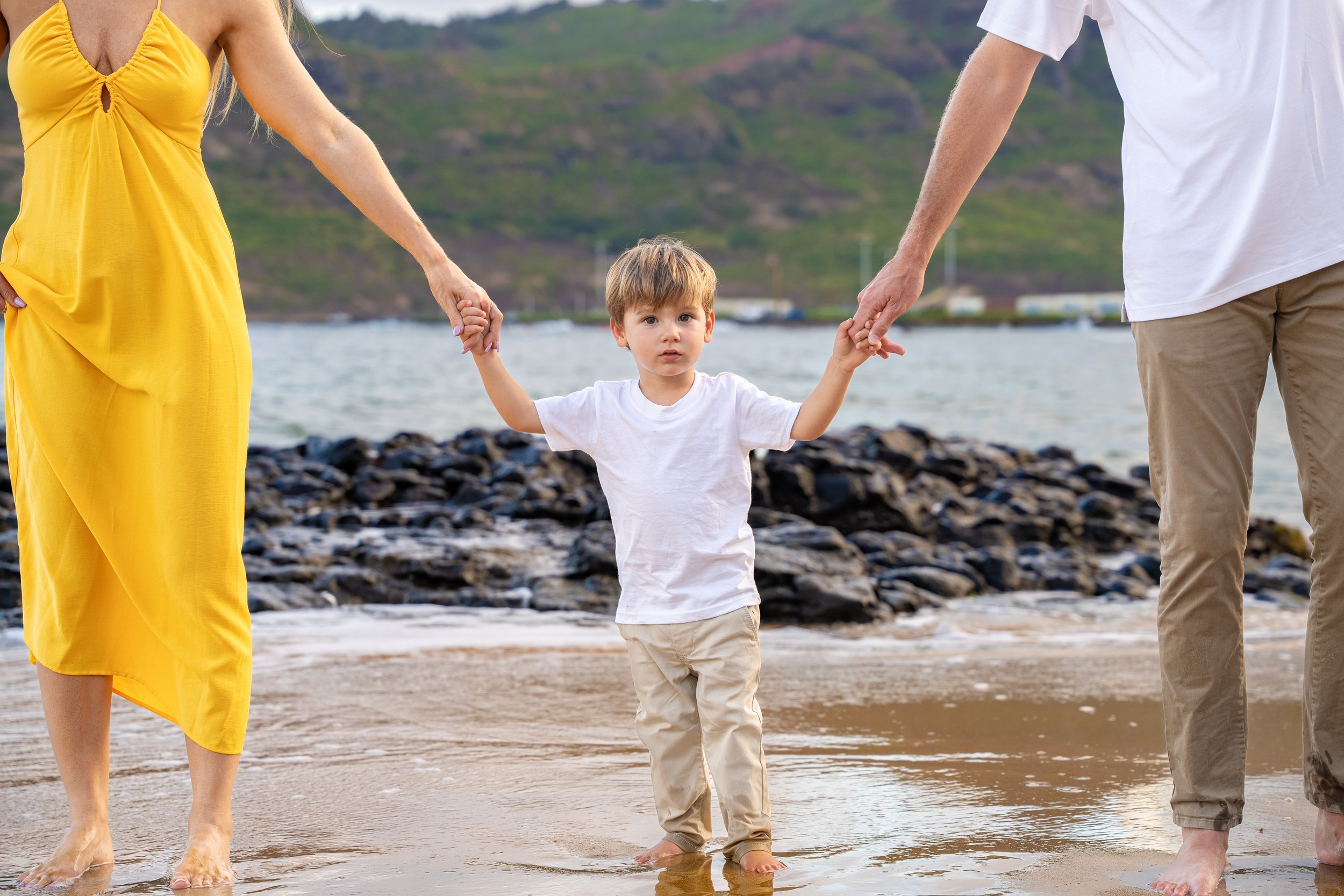Young boy holding his parents' hands while standing on a rocky Kauai beach.