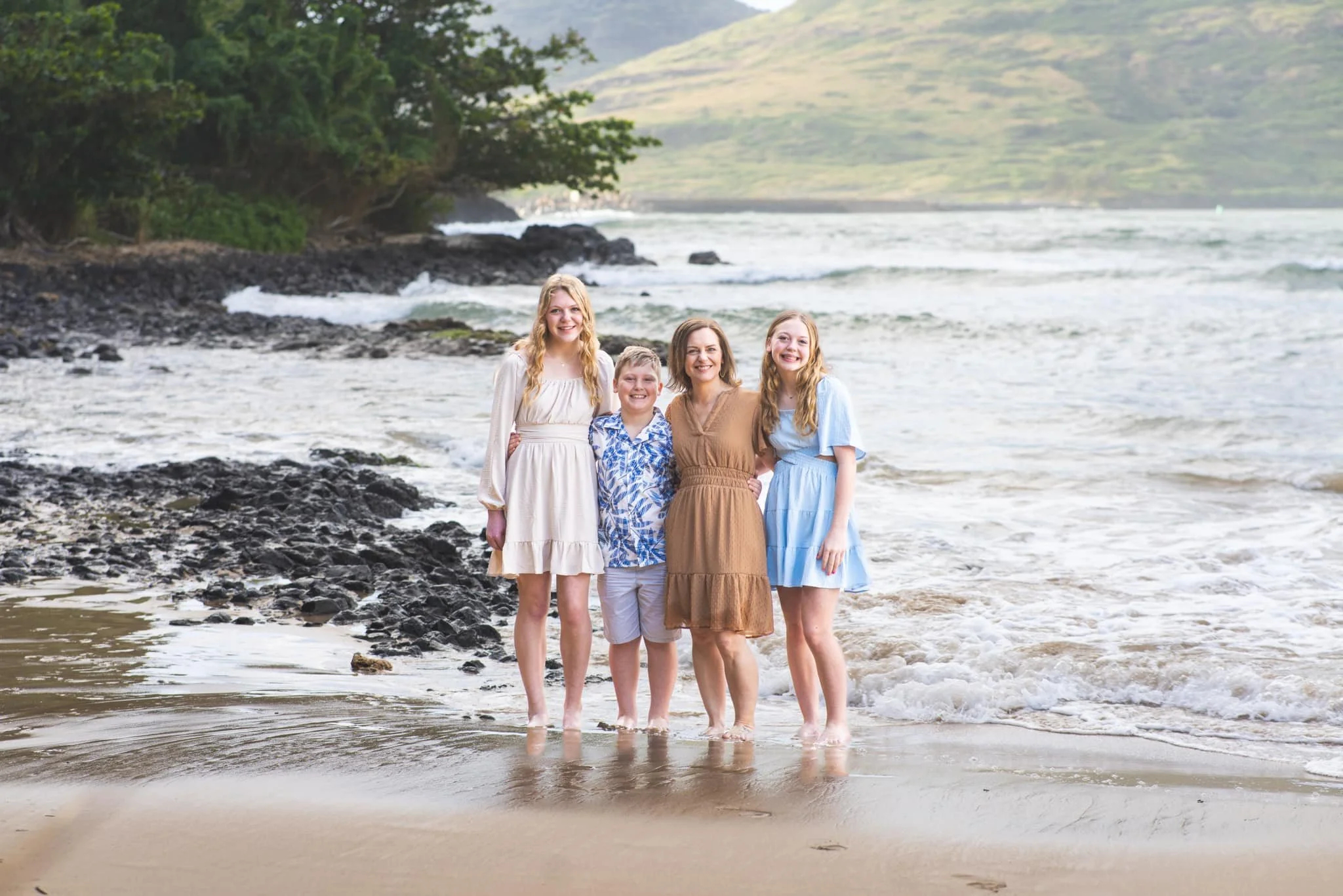 Mom with her three kids posing at the edge of the surf on a rocky Kauai beach.