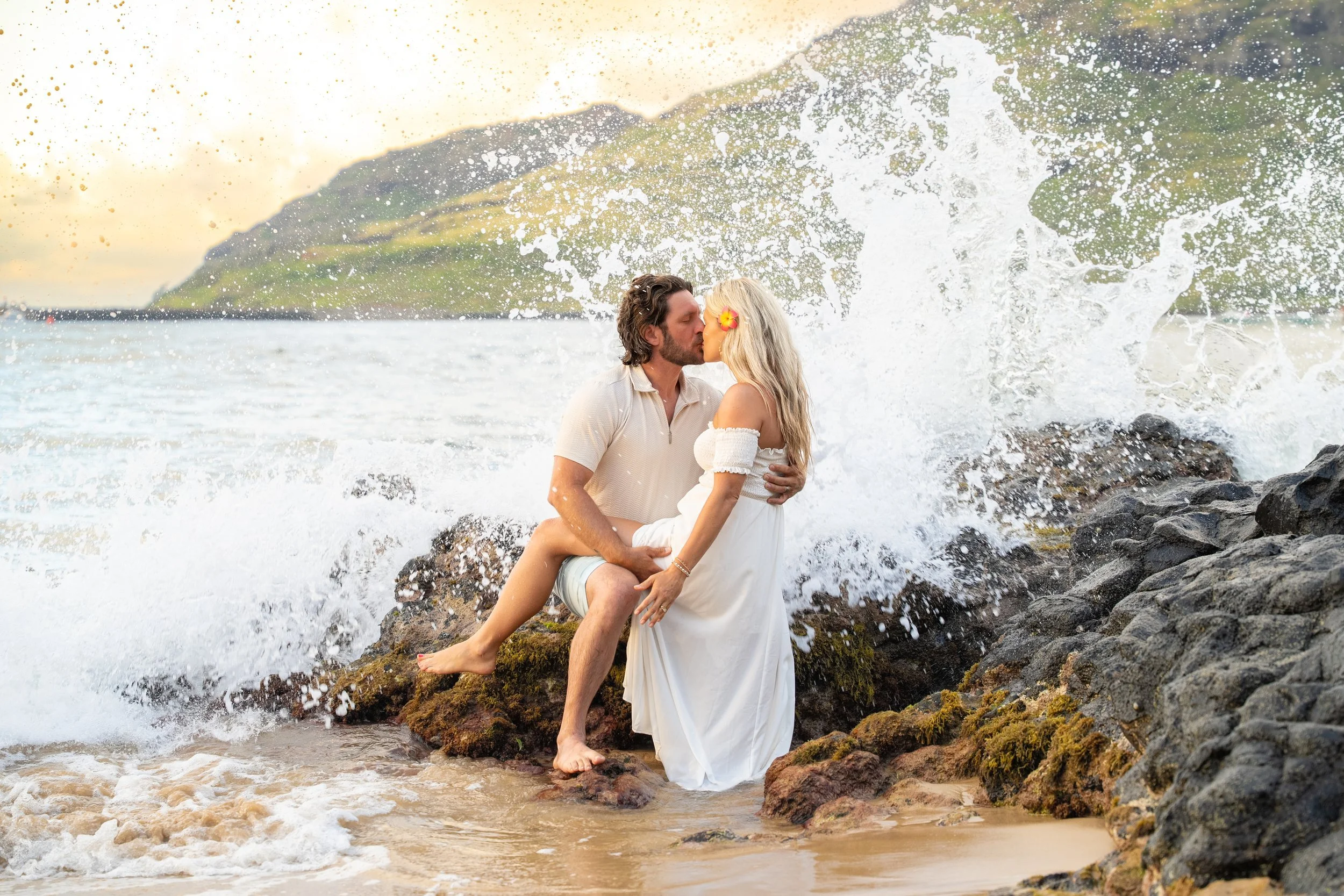 Couple sharing a kiss on a rocky Kauai shore as a wave splashes behind them.