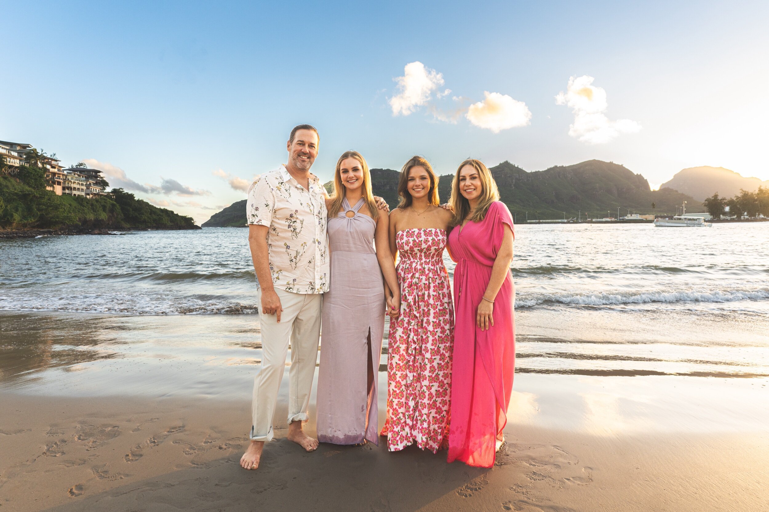 Family of four poses on the shoreline at sunset with Kauai mountains glowing behind them.