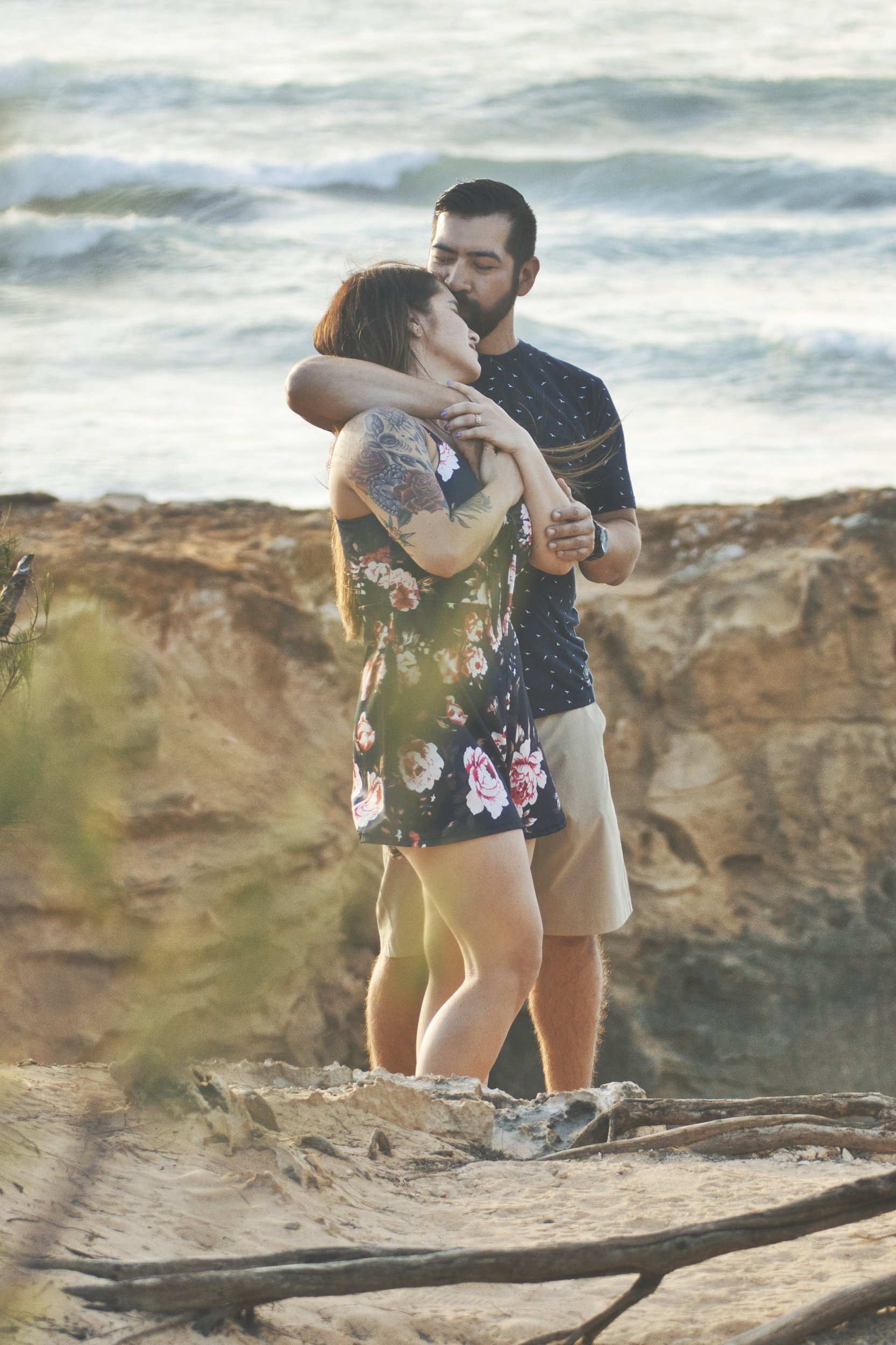 Couple embracing on the rocky shore with waves behind them at Shipwreck Beach, Kauai.