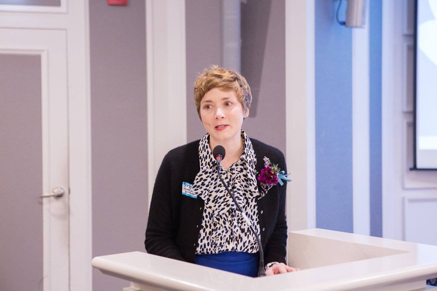 Alumni Affairs Director, Jane Nuzzo, is delivering her speech to the audience. She is wearing a black cardigan and has a tiny flower bouquet in her pocket.
