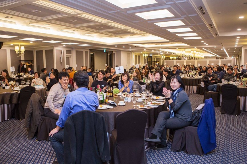 A panoramic view of the entire alumni party. Around 120 people are divided into groups of 10, sitting around round tables, engaging in conversations.