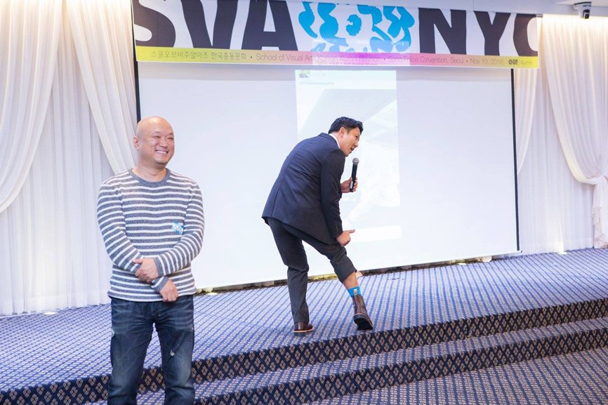 In front of the stage adorned with an SVA NYC banner, there is a man wearing a black suit holding a microphone. He lifts the hem of his pants, revealing sky blue socks. To his left, there is a man wearing a checked shirt, smiling.