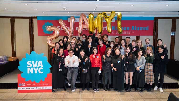 Twenty eight men and women are posing on the stage. In the background, there is a red banner that reads "School of Visual Arts Korean Alumni," and in front of it, there is a blue-red combination signboard with white letters that says "SVANYC." On top