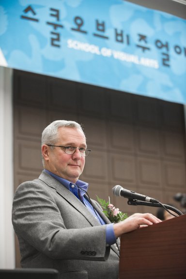The Vice President of SVA, A. Rhodes, is giving a speech at the podium. There is an SVA Alumni Association banner hanging behind them.
