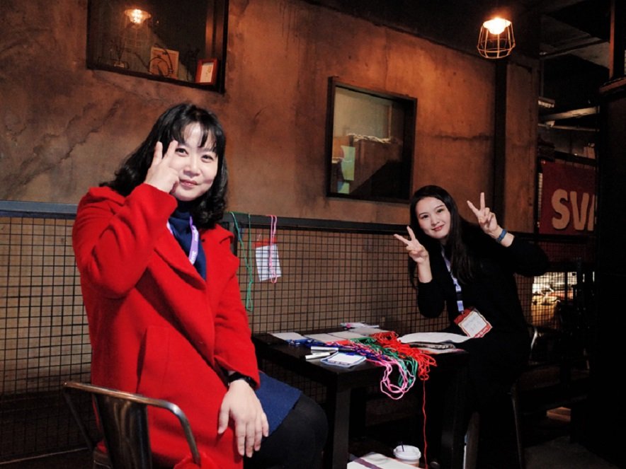 On the left side, there is a woman wearing a red coat, and she is making a V sign. On the right side, there is a seated woman who is also making a V sign with both of her hands.