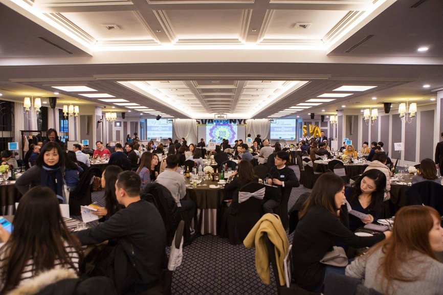 Another panoramic view of the entire alumni party. Roughly 120 attendees are organized into clusters of ten, positioned around circular tables, immersed in dialogue and exchanges.