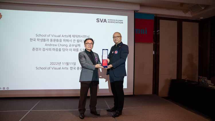 Former alumni association president Kim Takhoon and Andrew Chang are posing on the stage while holding plaques. In the background, there is a banner that reads: "To Andrew Chang, who has been a great support for Korean students and alumni while worki