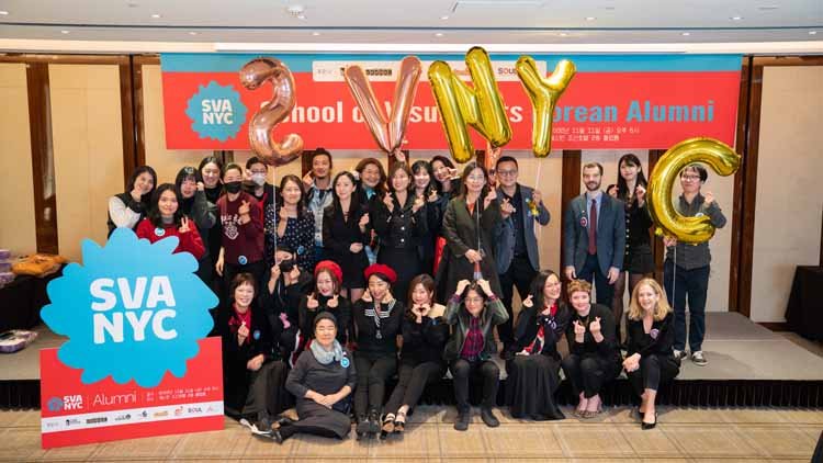 Twenty nine men and women are posing on the stage. In the background, there is a red banner that reads "School of Visual Arts Korean Alumni," and in front of it, there is a blue-red combination signboard with white letters that says "SVANYC." On top