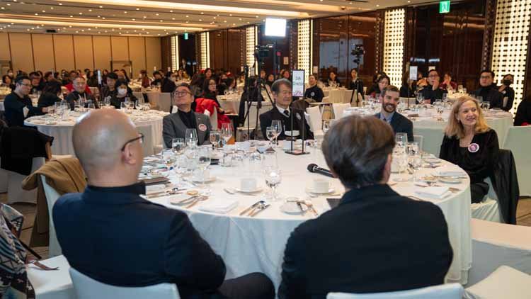 Here is the overall view of the alumni event. Multiple white round tables are placed, and around each table, there are approximately nine alumni seated.