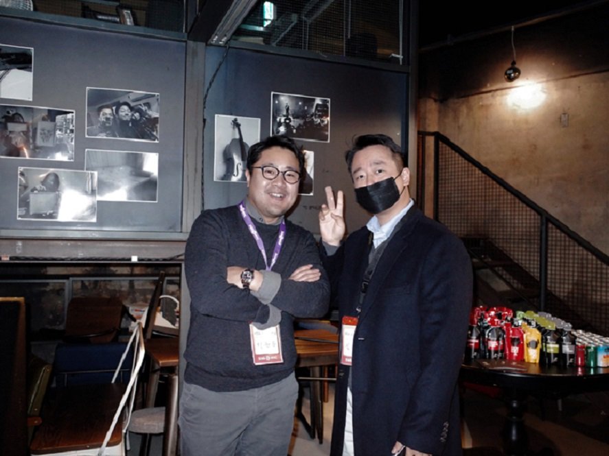 Here is a description of the scene: In the event hall, two men are posing. The man on the left has his arms crossed, while the man on the right is making a V sign with his fingers.