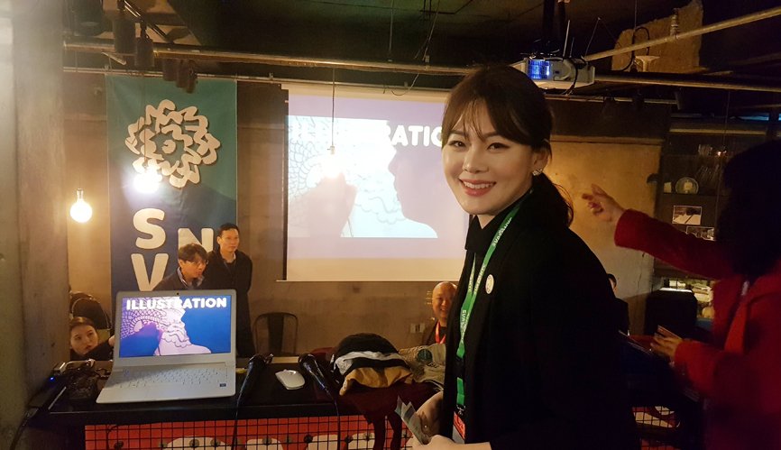 Alumni MC Kim Yeonji is looking into the camera. Behind her, there is a projector screen, and on the left side, the SVA NYC banner is hanging down.