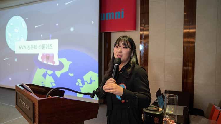 On the podium, there is a female wearing a black outfit, giving a speech. On the left side, there is a screen playing an animation.