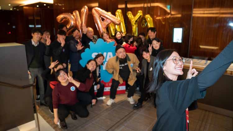 There are 15 males and females gathered around a blue signboard that says "SVA NYC," striking poses. The female on the far right in the front is holding up a camera to take a selfie.