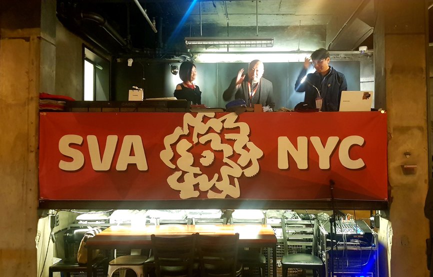 From the stage located on the second floor, Alumni Association President Kim Takhoon is giving a speech. On either side of the stage, there are men and women standing. A banner with "SVA NYC" written on it is displayed on the podium.