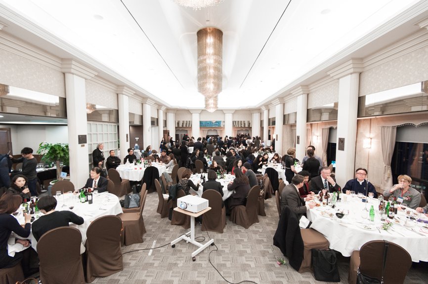 A panoramic view of the entire alumni party. Around 120 people are divided into groups of 10, sitting around round tables, engaging in conversations.
