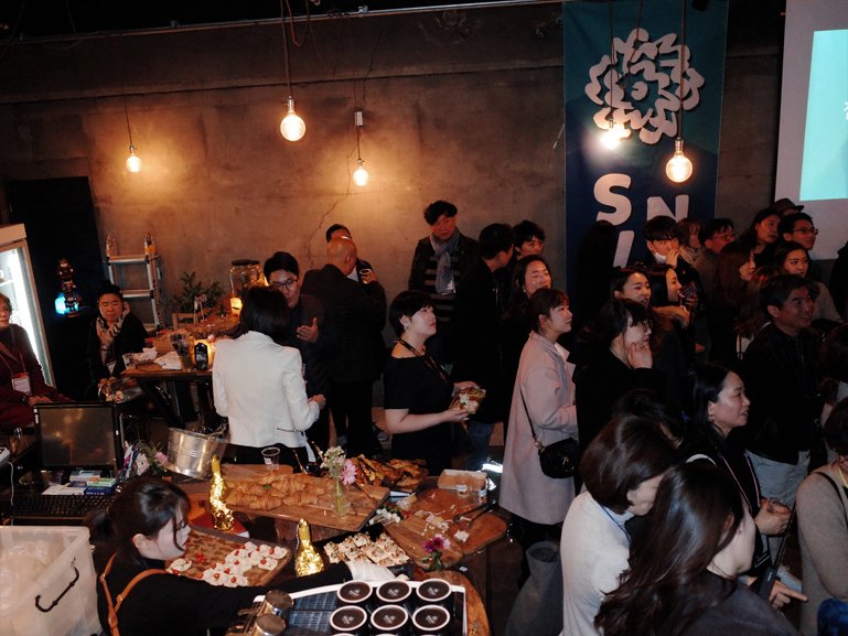 In the event hall, around 20 graduates are facing towards the right side. On the lower left corner, a buffet is set up, and at the back on the right side, there is a blue SVA NYC banner displayed.