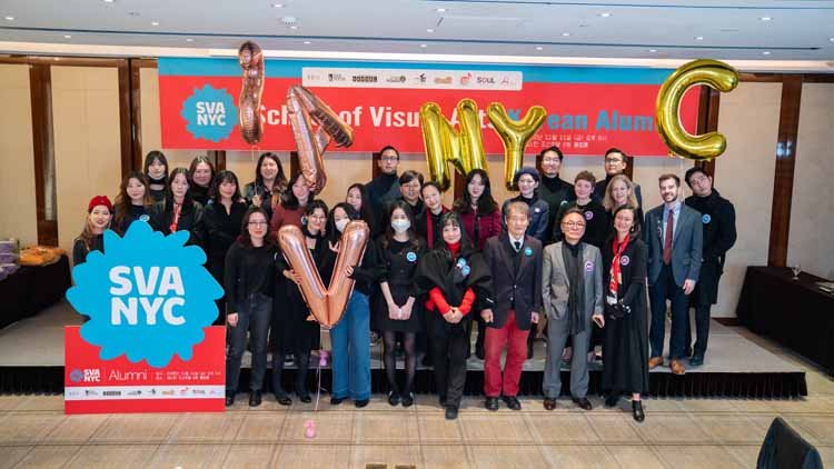 Twenty nine men and women are posing on the stage. In the background, there is a red banner that reads "School of Visual Arts Korean Alumni," and in front of it, there is a blue-red combination signboard with white letters that says "SVANYC." On top