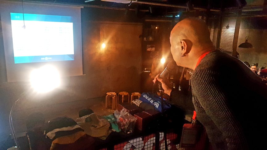 A man is holding a microphone and facing a projector while speaking. In front of him, there are displayed products.