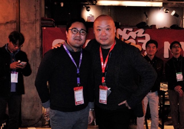 Two men wearing black clothes are posing with their hands on each other's shoulders.
