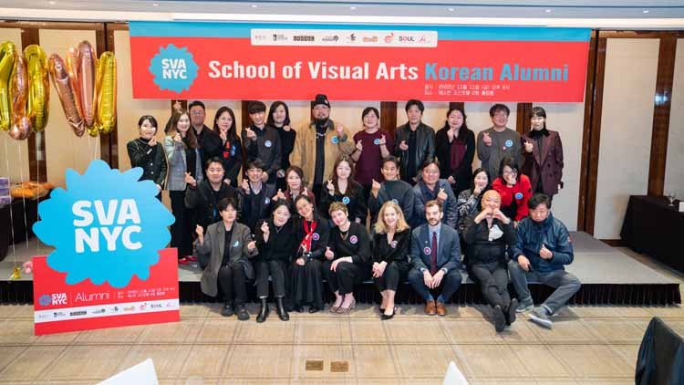 Twenty eight men and women are posing on the stage. In the background, there is a red banner that reads "School of Visual Arts Korean Alumni," and in front of it, there is a blue-red combination signboard with white letters that says "SVANYC." On top