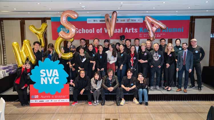 Twenty five men and women are posing on the stage. In the background, there is a red banner that reads "School of Visual Arts Korean Alumni," and in front of it, there is a blue-red combination signboard with white letters that says "SVANYC." On top