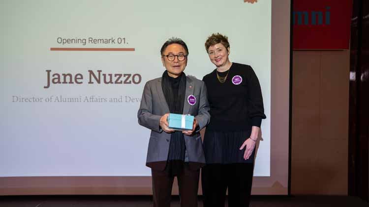 Andrew Chang, holding a mint-colored box, and Jane Nuzzo, the Alumni Affairs Director, are posing together on the stage. The screen displays "Alumni Affairs Director Jane Nuzzo."