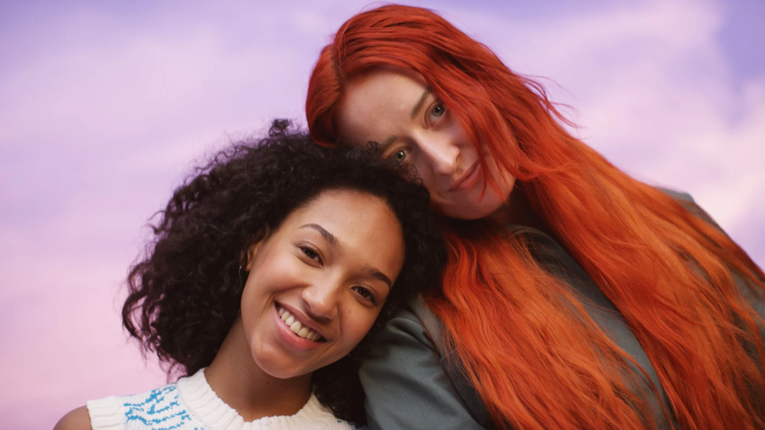 Two women smiling and leaning on each other against a pink and purple sky background, one with curly dark hair and the other with long red hair.