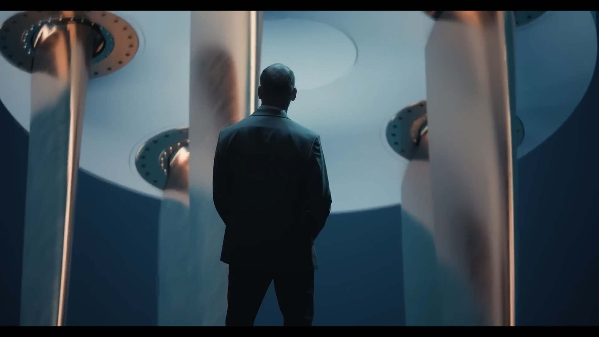 A man in a suit standing with his back to the camera, looking at a large, modern art installation or architectural structure with metallic and circular elements.