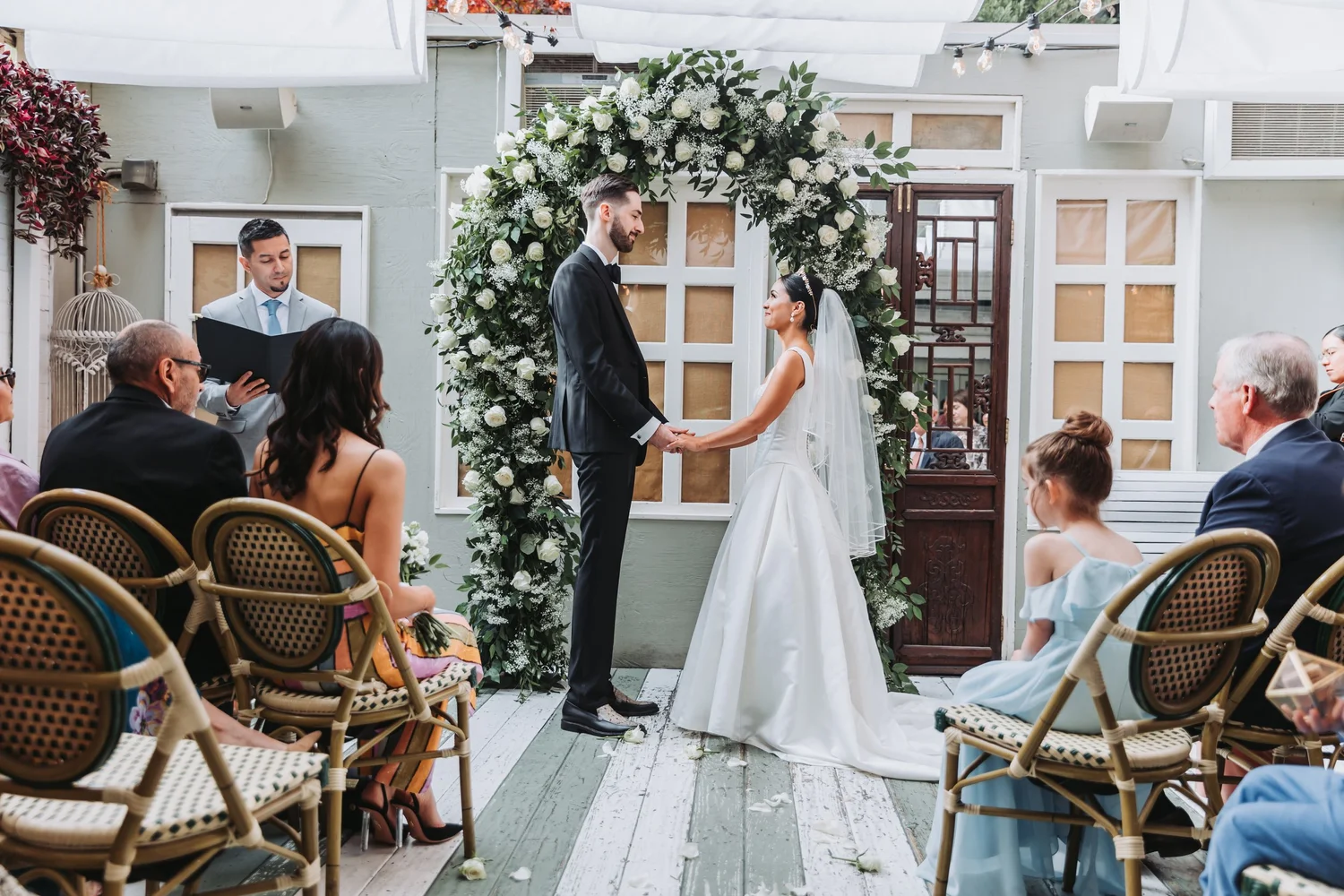 Bride and groom exchange vows beneath a floral arch at an outdoor ceremony.