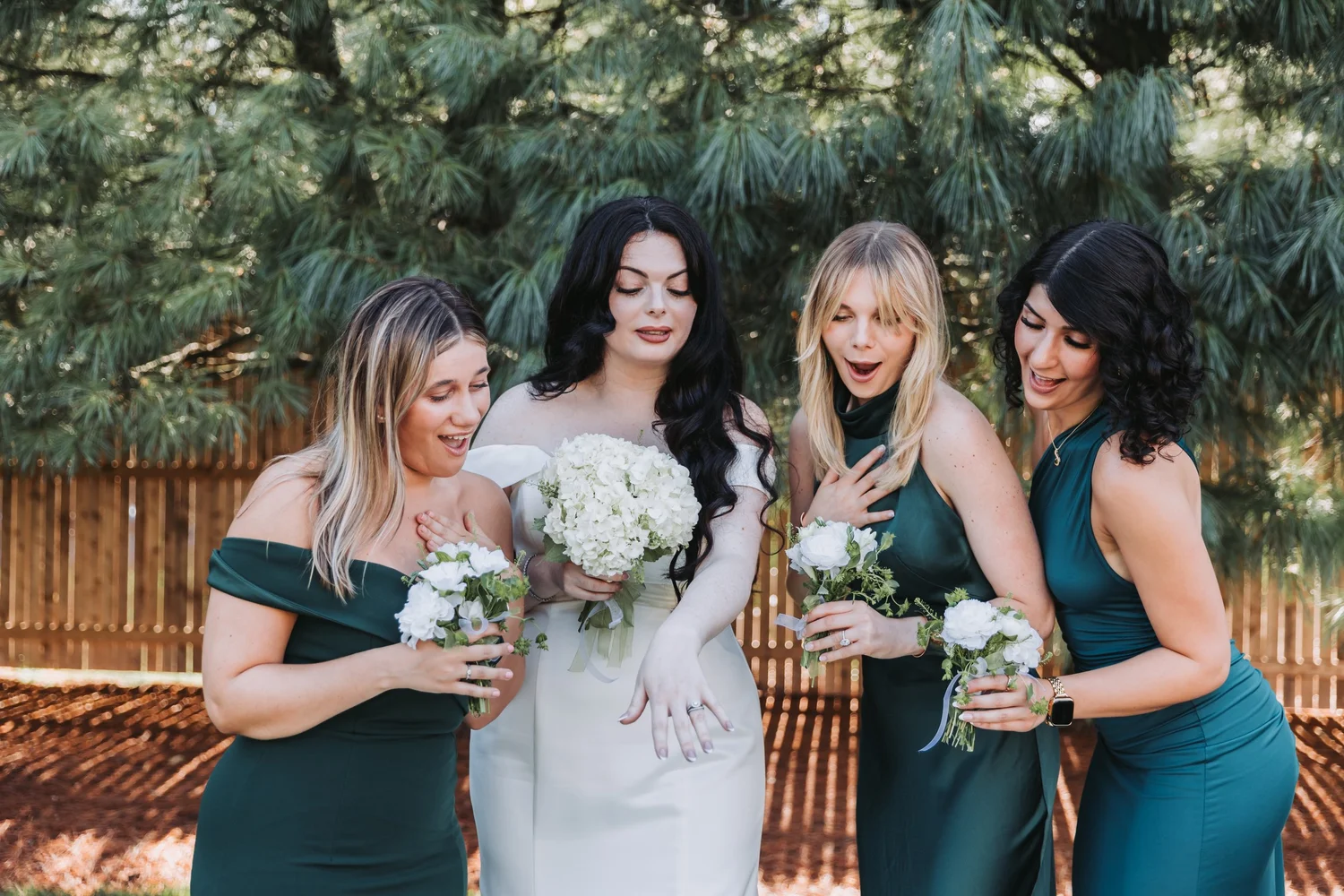 Bride holding her bouquet among bridesmaids in teal dresses beneath leafy trees.