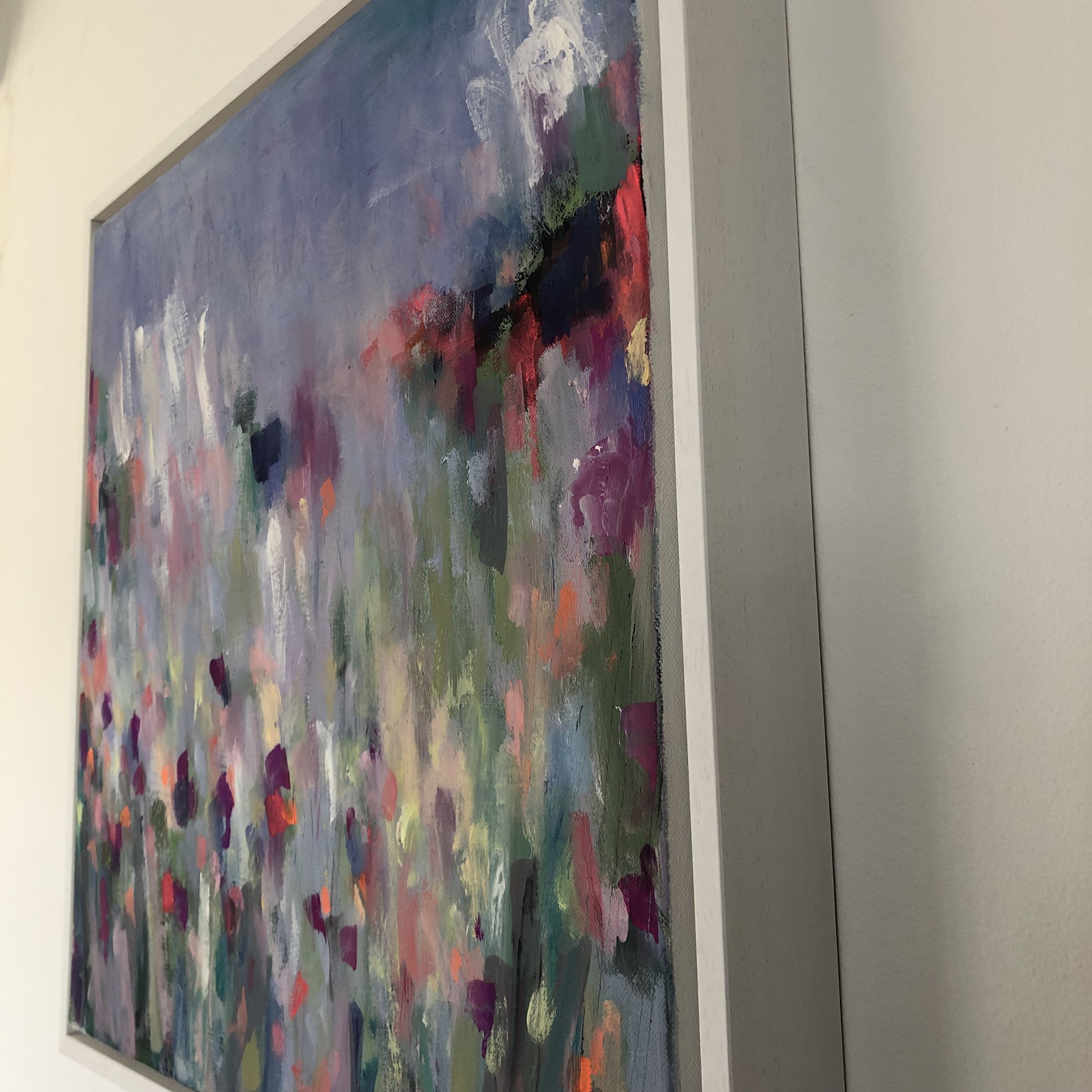 Side view of painting on a wall in a white tray frame. Vibrant abstract painting showing variety of colours, based on a farm’s driveway covered in wildflowers here in north Wales.
