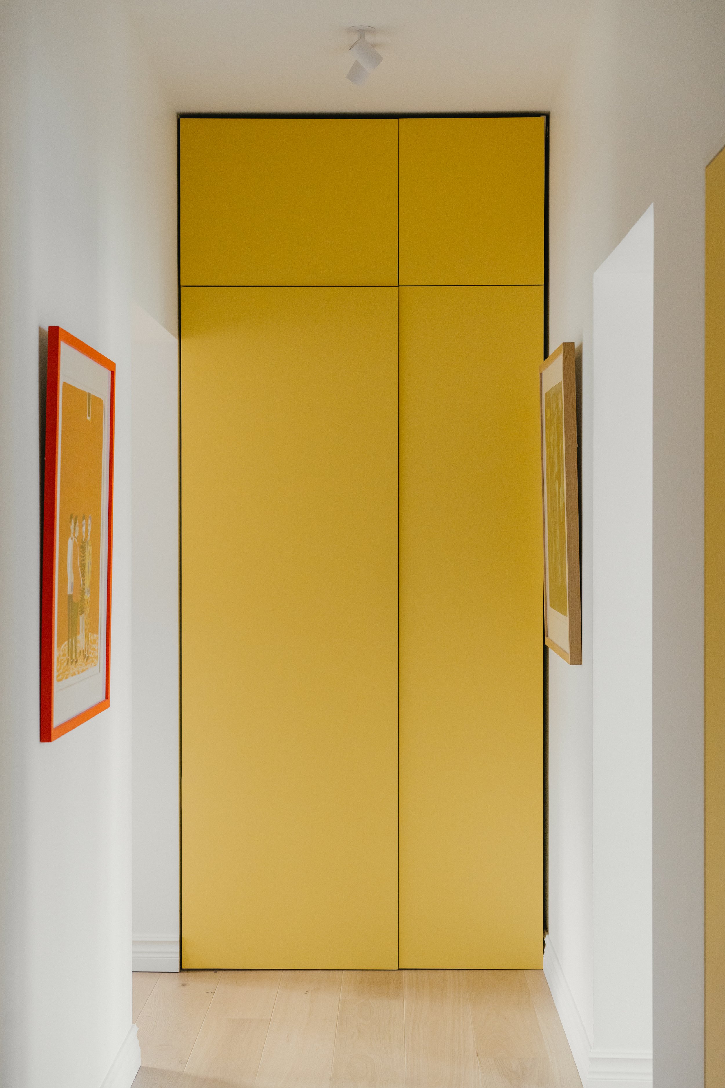 A hallway with white walls and wood flooring, featuring a yellow closet with four doors and two framed artworks on the walls.