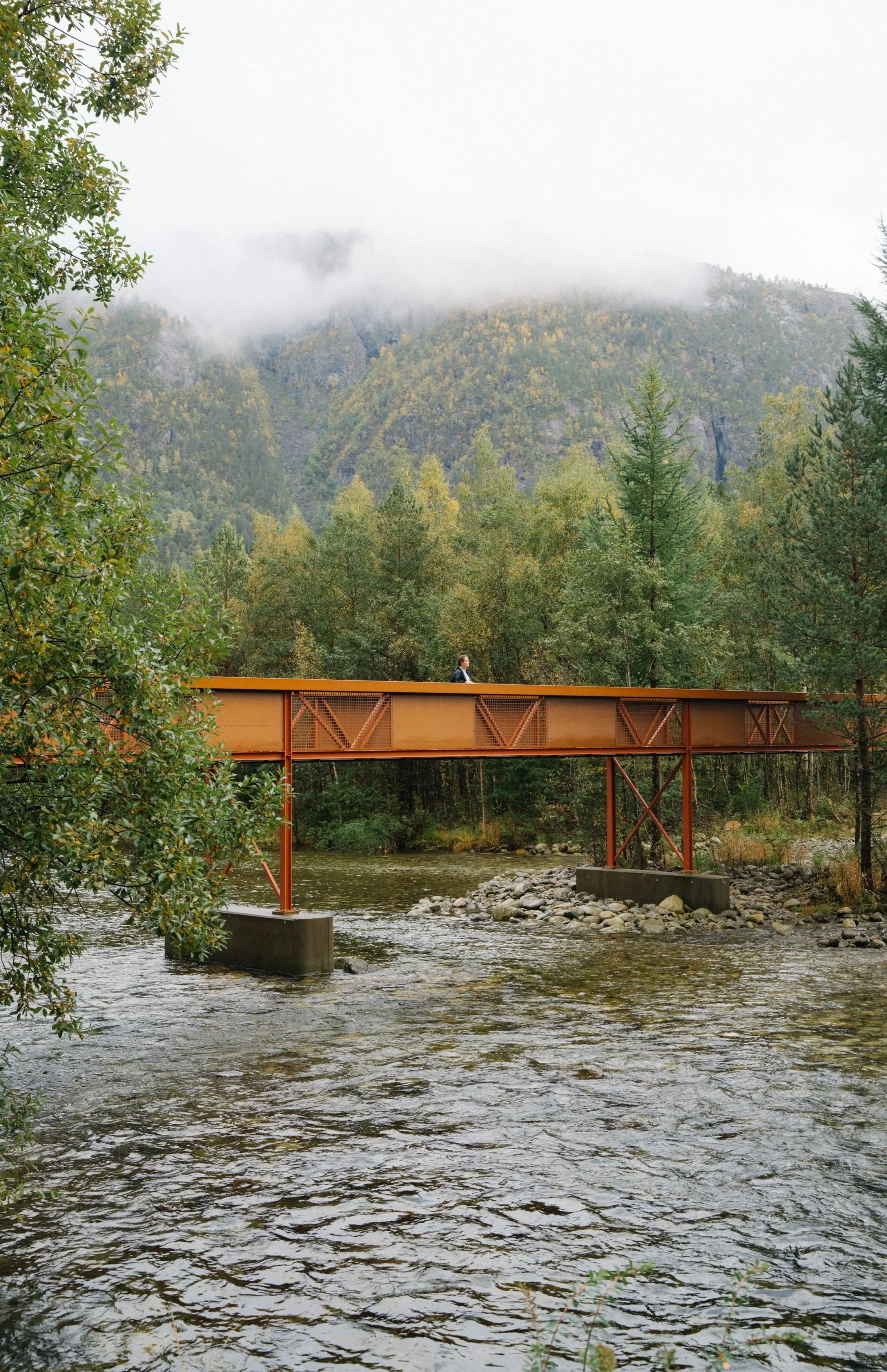 A person walking on a rusty orange metal bridge over a river in a forested area with mountains and low clouds in the background.