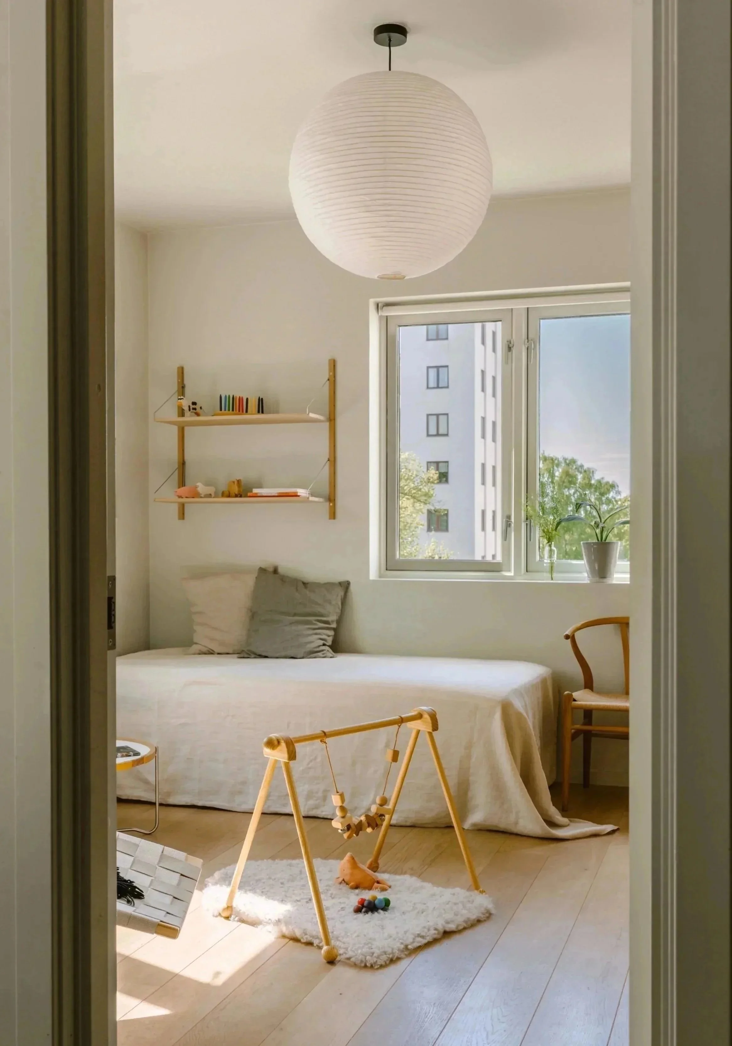 Minimalist bedroom with a single bed, a window with green plants, a wooden chair, a small plush carpet, a child's play gym, and a paper lantern ceiling light.