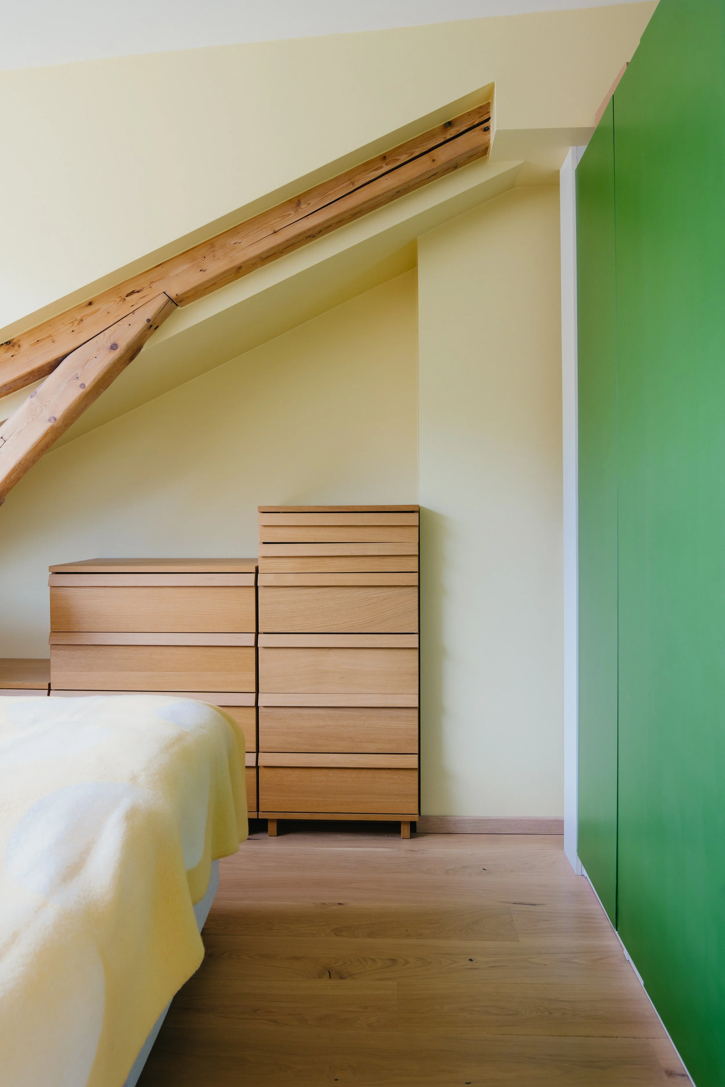 Interior view of a bedroom with wooden flooring, a yellow wall, a bed with yellow blankets, a wooden dresser, and a large green wardrobe, under a sloped ceiling with wooden beams.