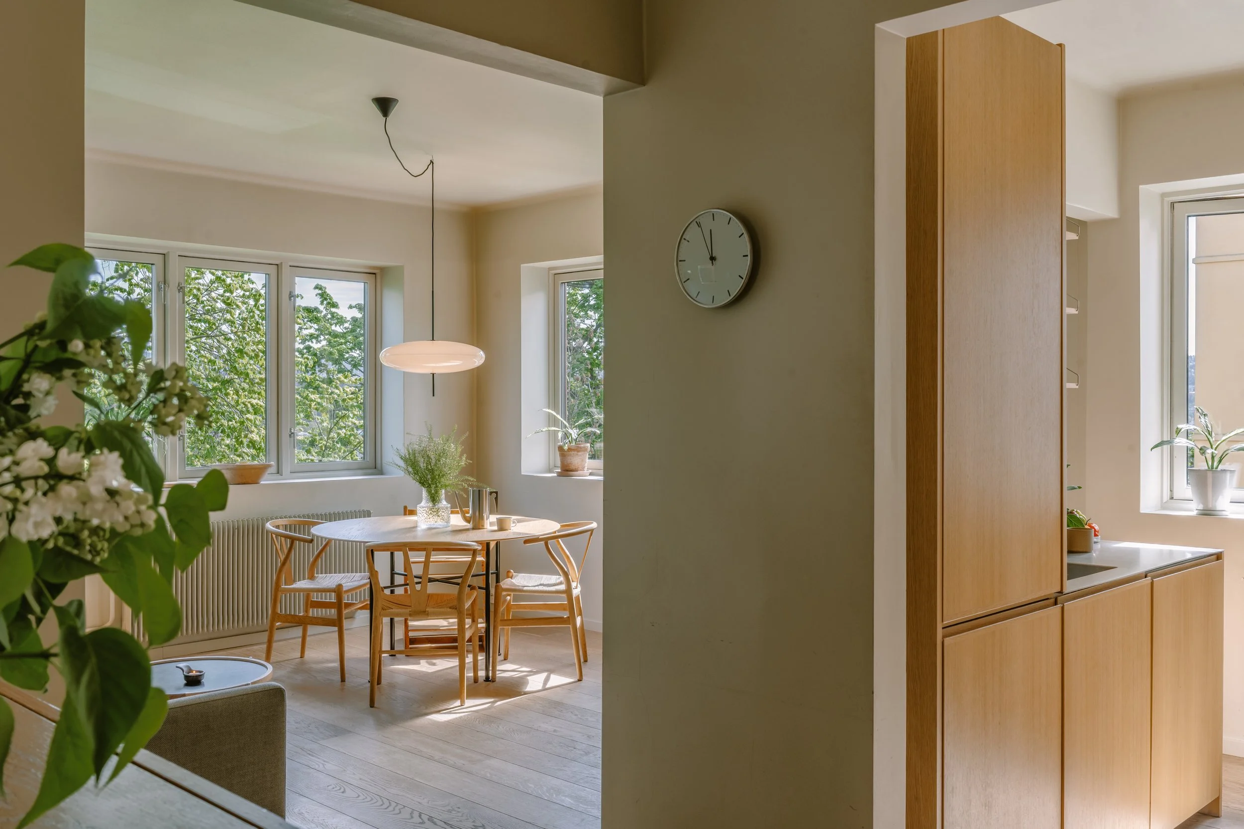 Bright dining area with a round wooden table and four chairs, large windows with green trees outside, potted plants on windowsills, a modern pendant light, and a clock on the wall.
