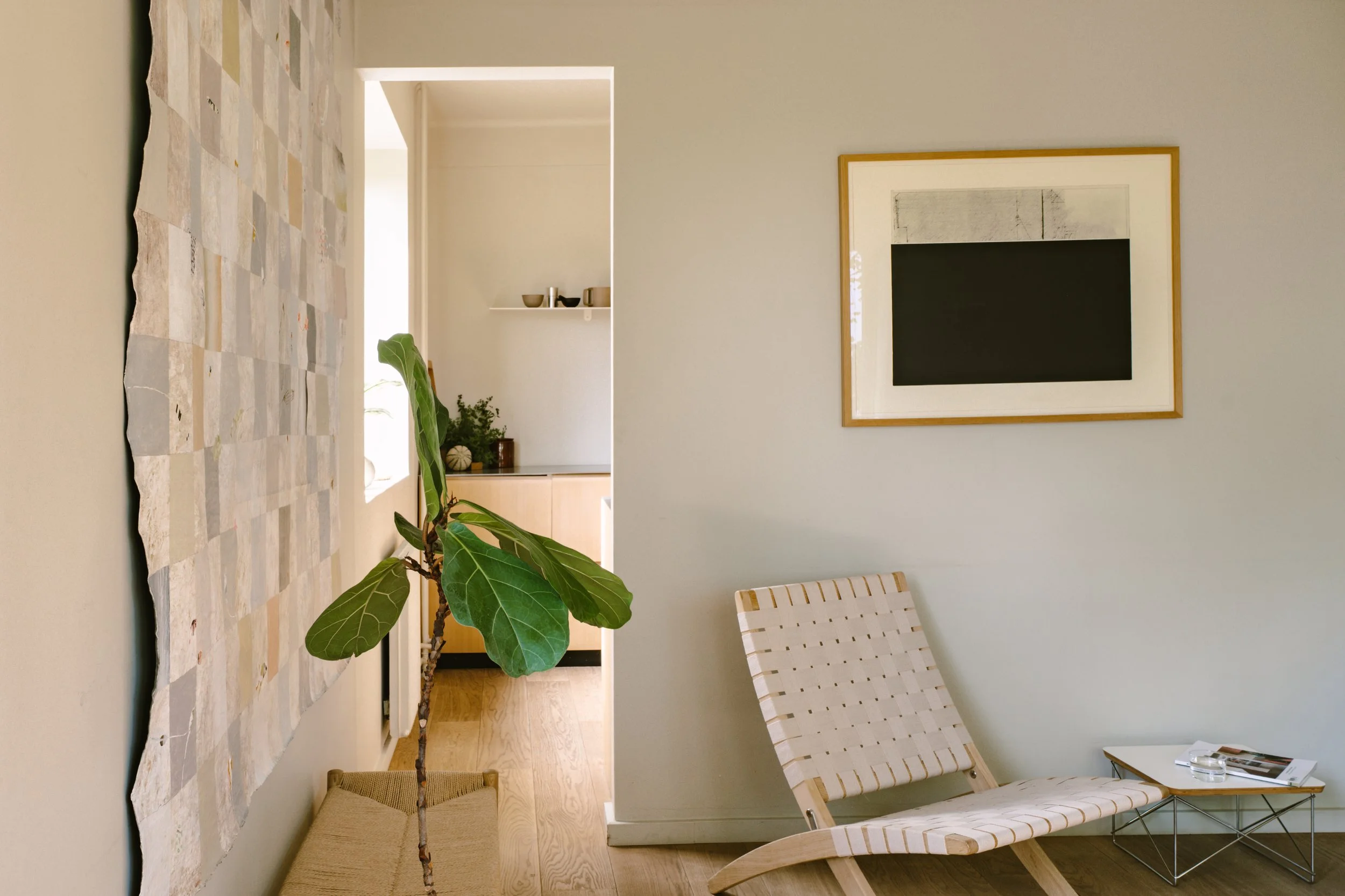Interior of a modern living room with a wicker lounge chair, side table with magazines, a potted plant, abstract art on the wall, and an open doorway leading to a kitchen area with shelves and decor.
